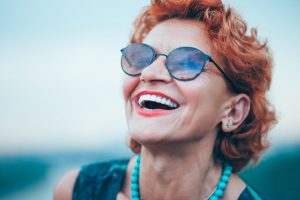 An older woman with red hair and sunglasses smiles, showing off her porcelain veneers.