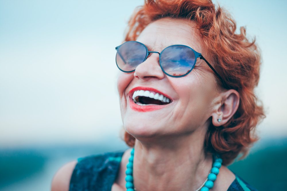Happy,Mature,Woman,Enjoying,Life An older woman with red hair and sunglasses smiles, showing off her porcelain veneers.